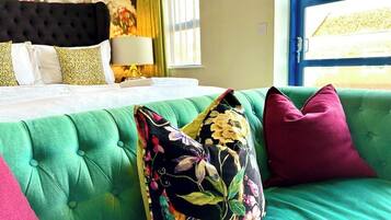Una habitación con un sofá tapizado en verde, una cama con ropa blanca y una almohada estampada con motivos florales.
