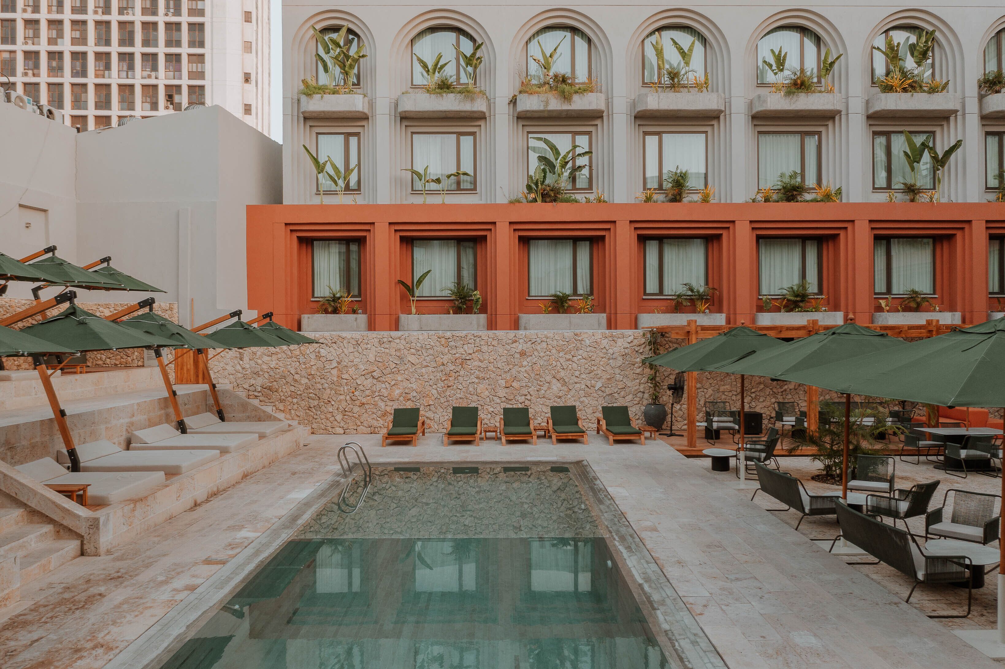 A pool area with lounge chairs and umbrellas, a stone wall, and a building with arched windows and plants.