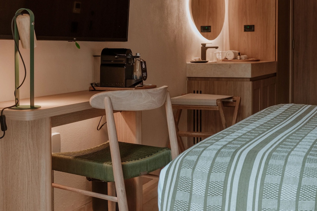 A compact hotel room with a bed, a desk with a chair, a coffee machine, and a large mirror.