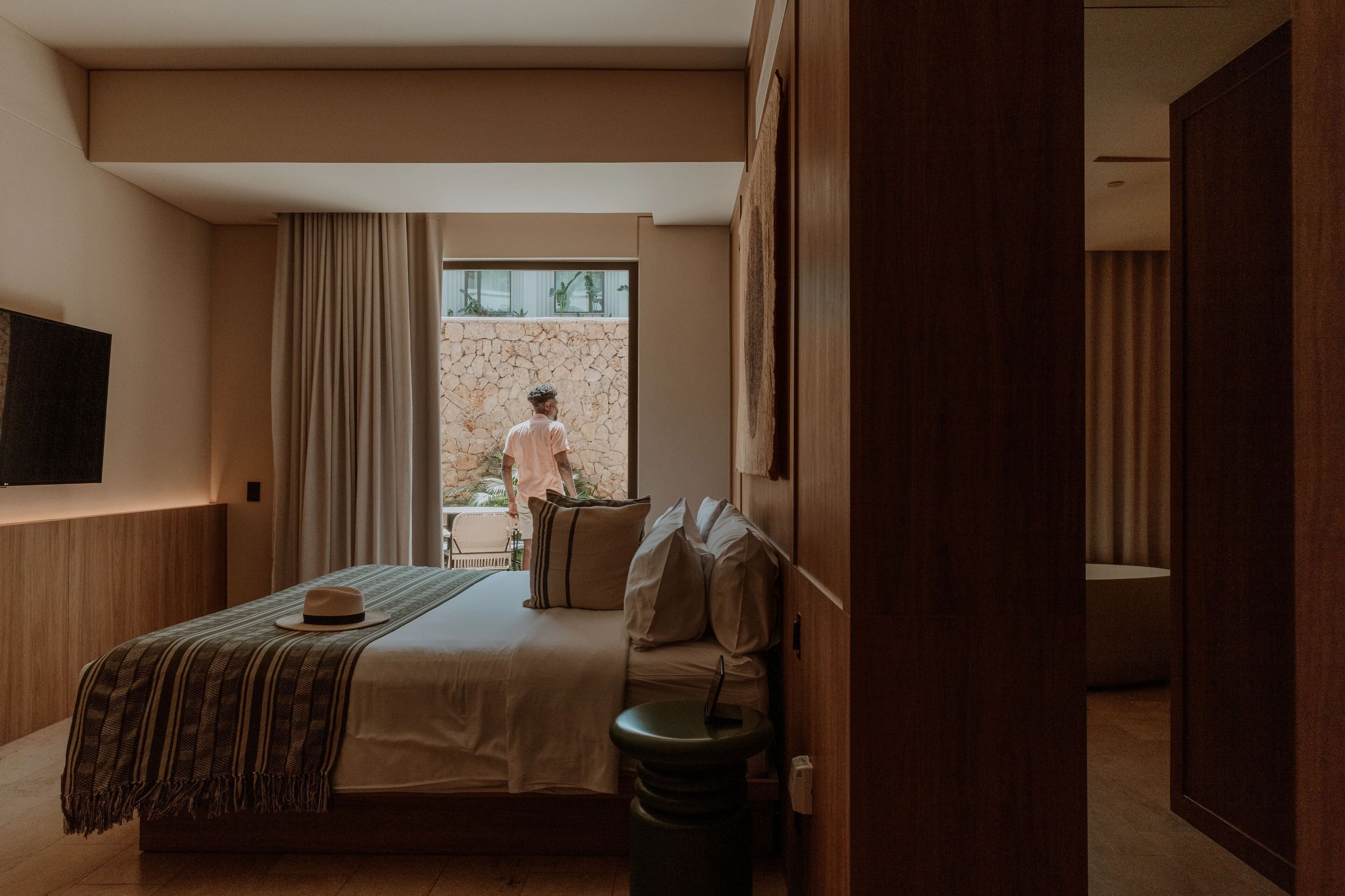 A modern hotel room with a large bed, a bedside table, a TV mounted on the wall, and a view of the outside balcony.