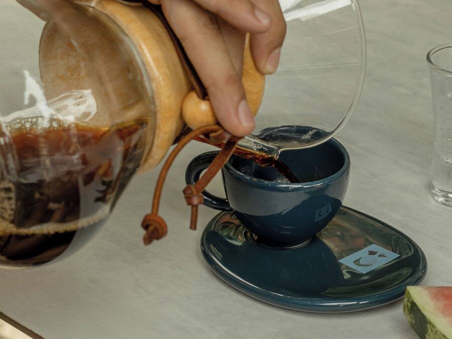 Une main verse un liquide sombre d’un récipient en bois dans une tasse en céramique bleue, accompagnée d’une soucoupe.