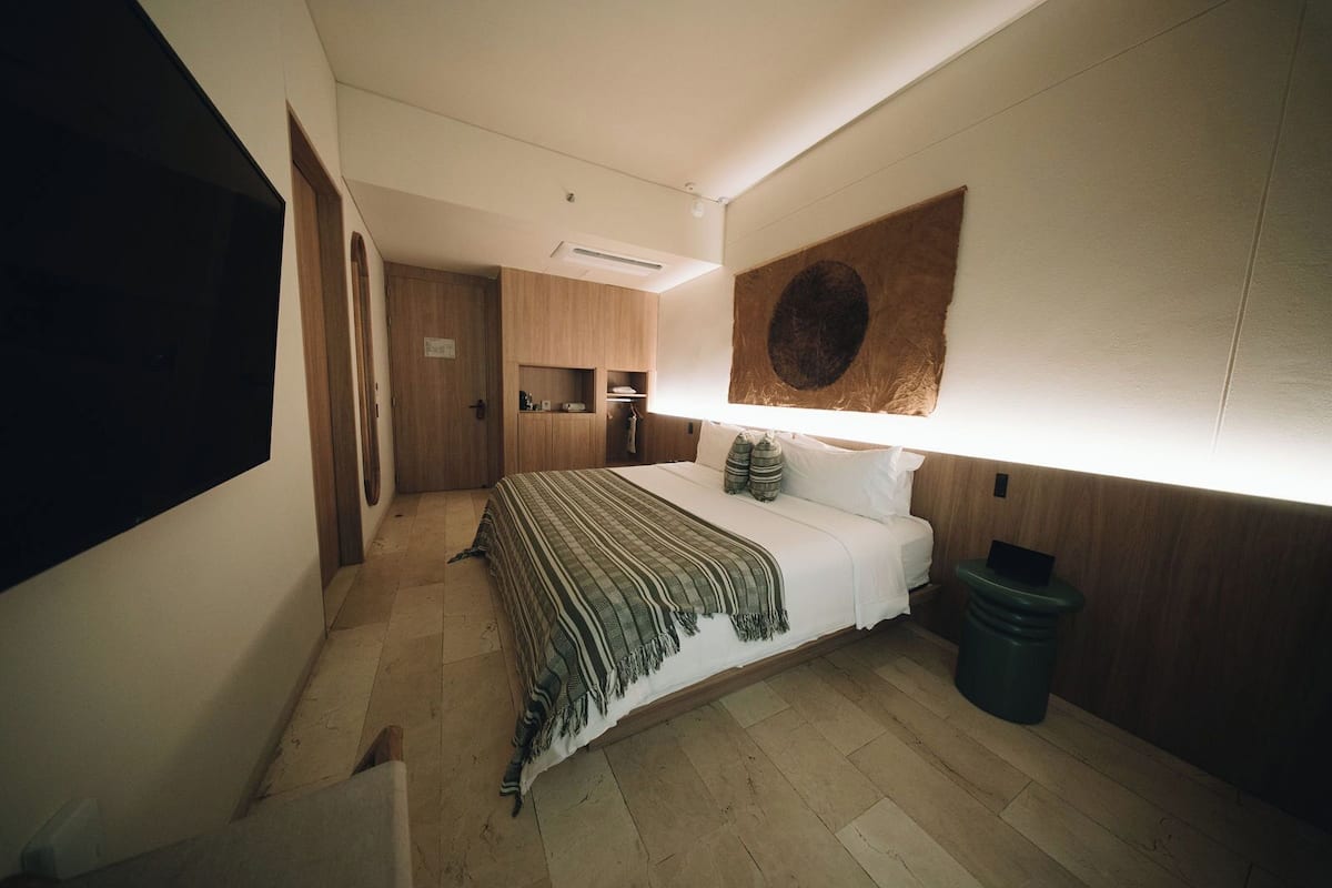 A bedroom with a bed, a wooden wardrobe, a small shelf, a wall-mounted TV, and a trash bin.