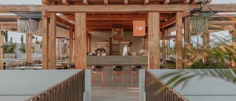 Un espace bar extérieur en bois, doté d’un comptoir et de tabourets, offrant une vue dégagée sur le bar intérieur, et un chemin menant à l’espace détente.