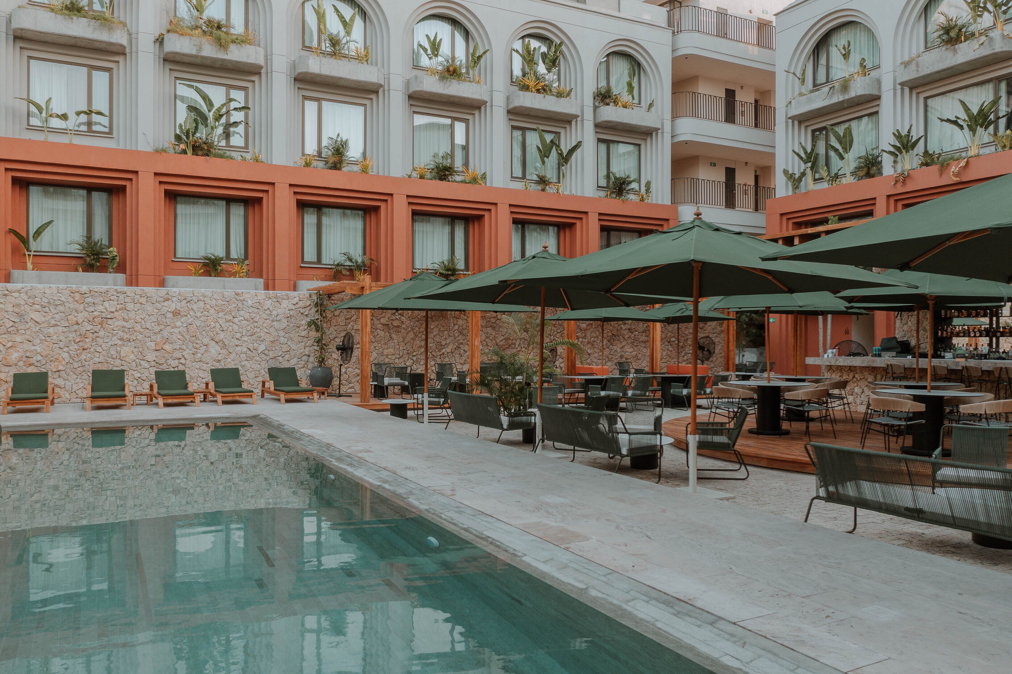A pool area with lounge chairs and umbrellas, surrounded by a stone wall and a building with arched windows and balconies.