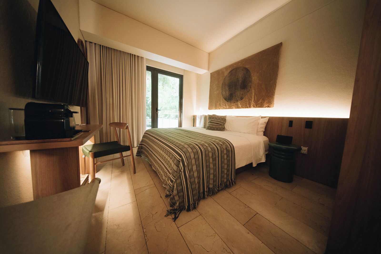 A hotel room with a bed, a desk with a chair, a large window, and a painting on the wall.