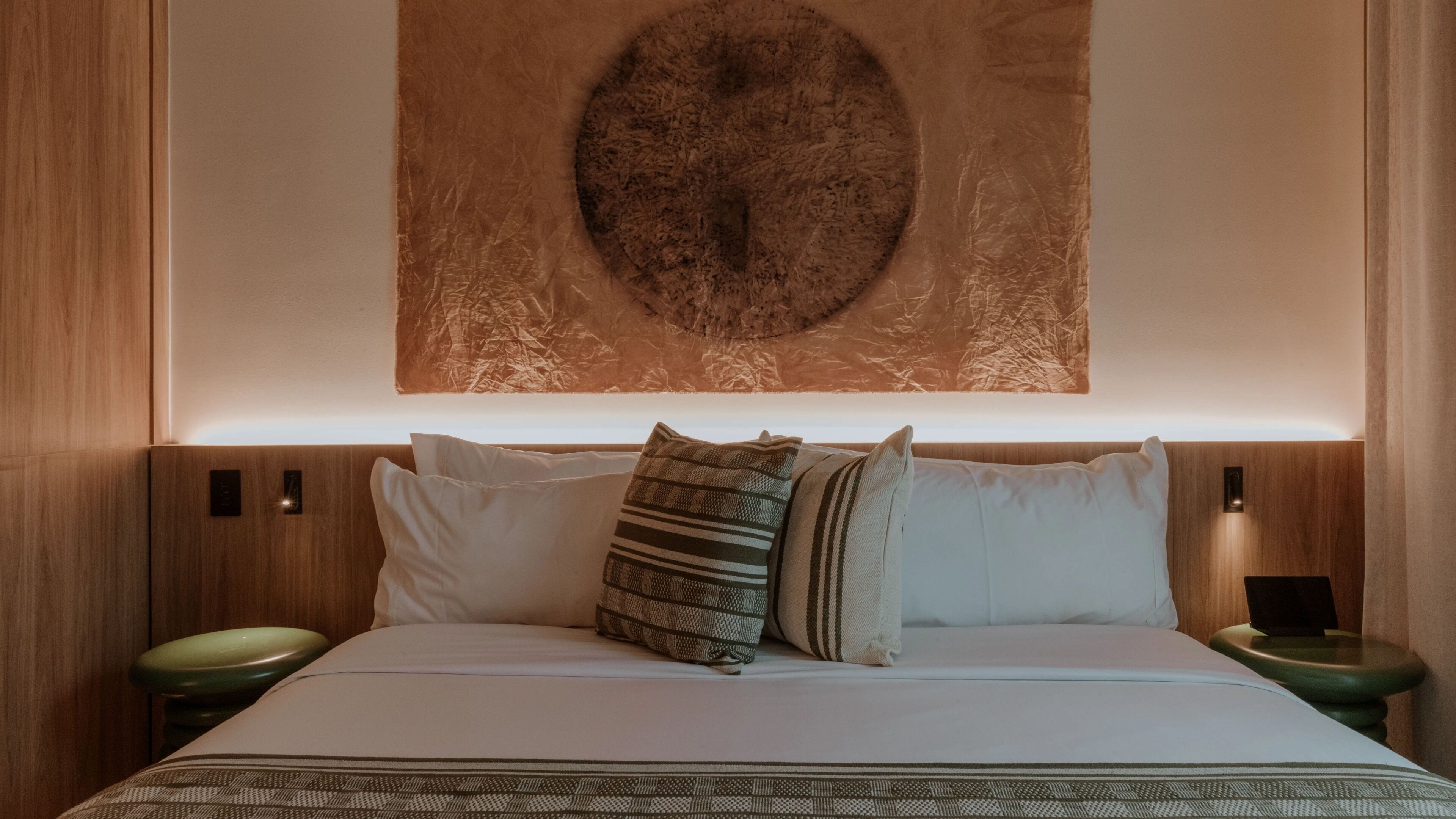 A neatly made bed with white linens and multiple pillows, including a striped one. There are two bedside tables with lamps, and a large circular artwork hangs above the bed.