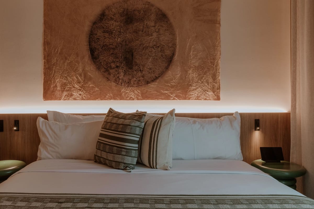 A neatly made bed with white linens and multiple pillows, including a striped one. There are two bedside tables with lamps, and a large circular artwork hangs above the bed.