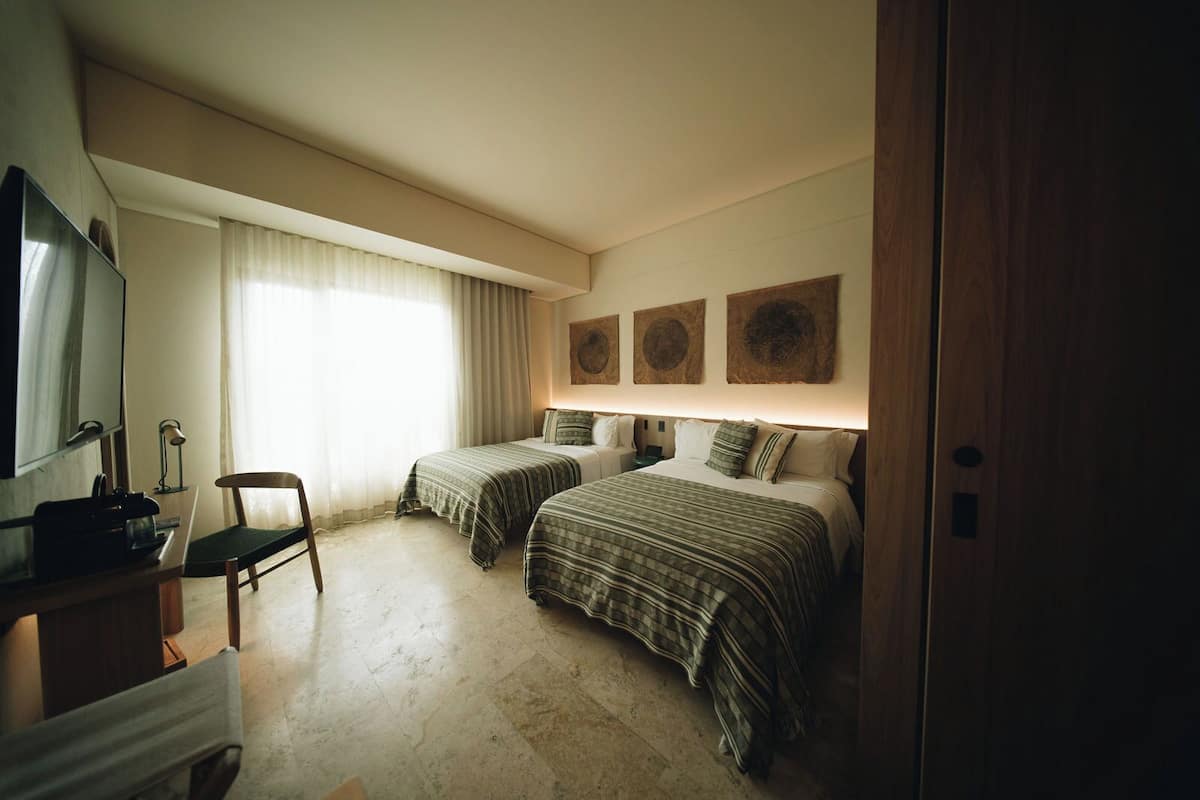 A hotel room with two beds, a desk, a chair, a TV, and a window with curtains.