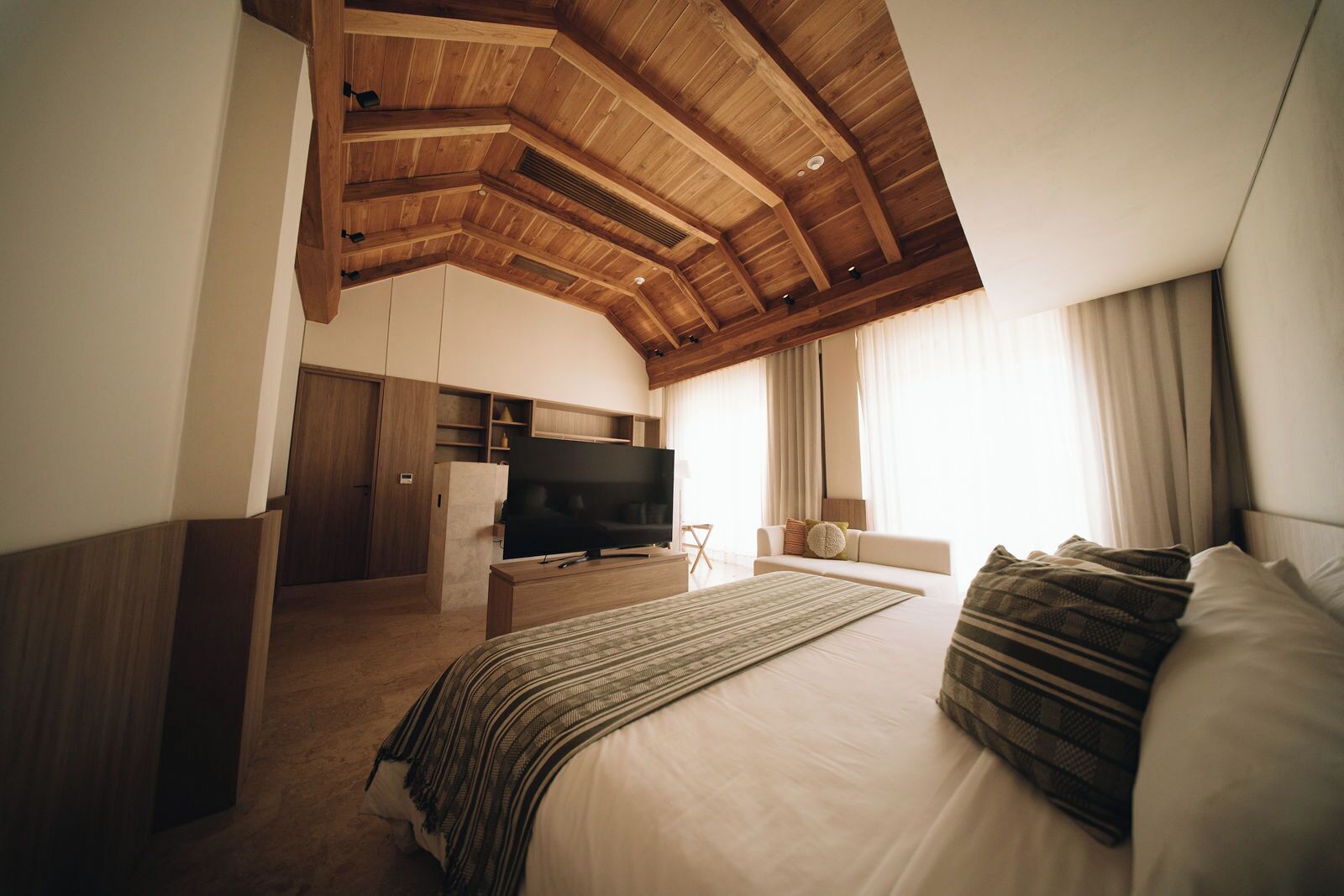 A bedroom with a large bed, a TV, wooden ceiling beams, and a window with curtains.