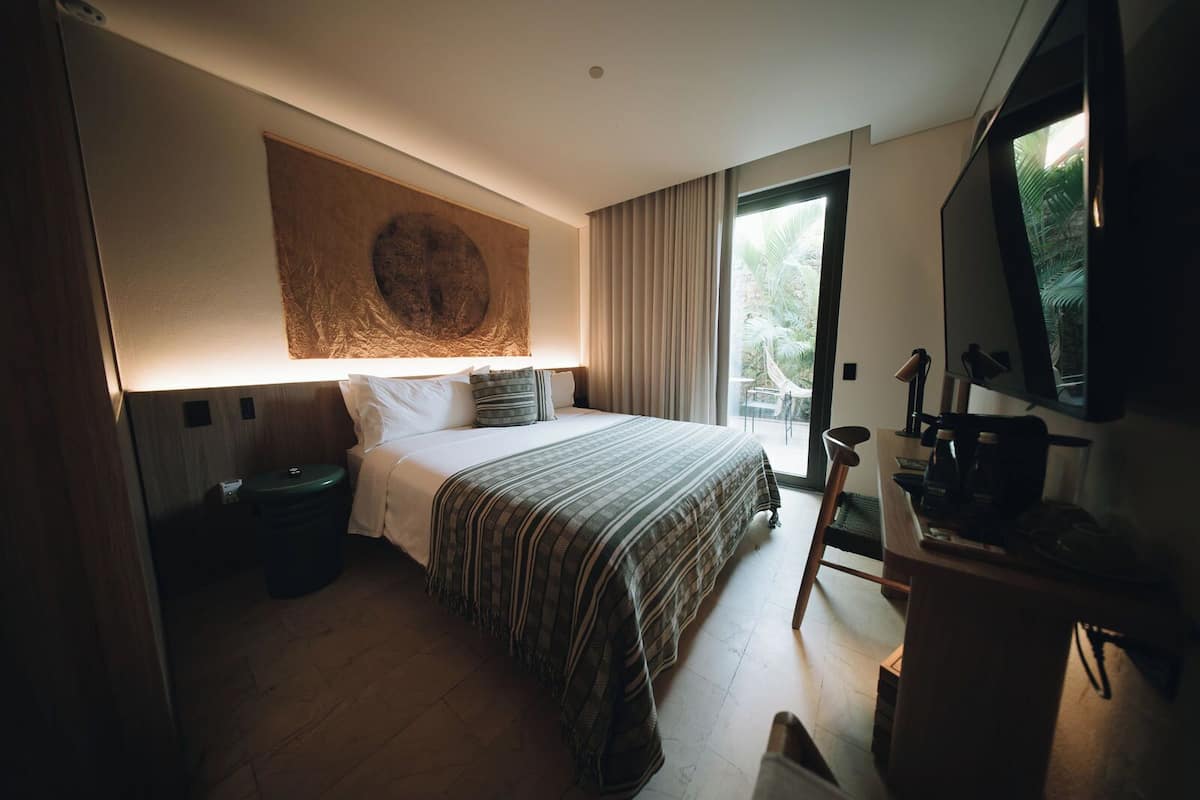 A hotel room with a bed, a desk with a chair, a TV, and a large window with curtains.