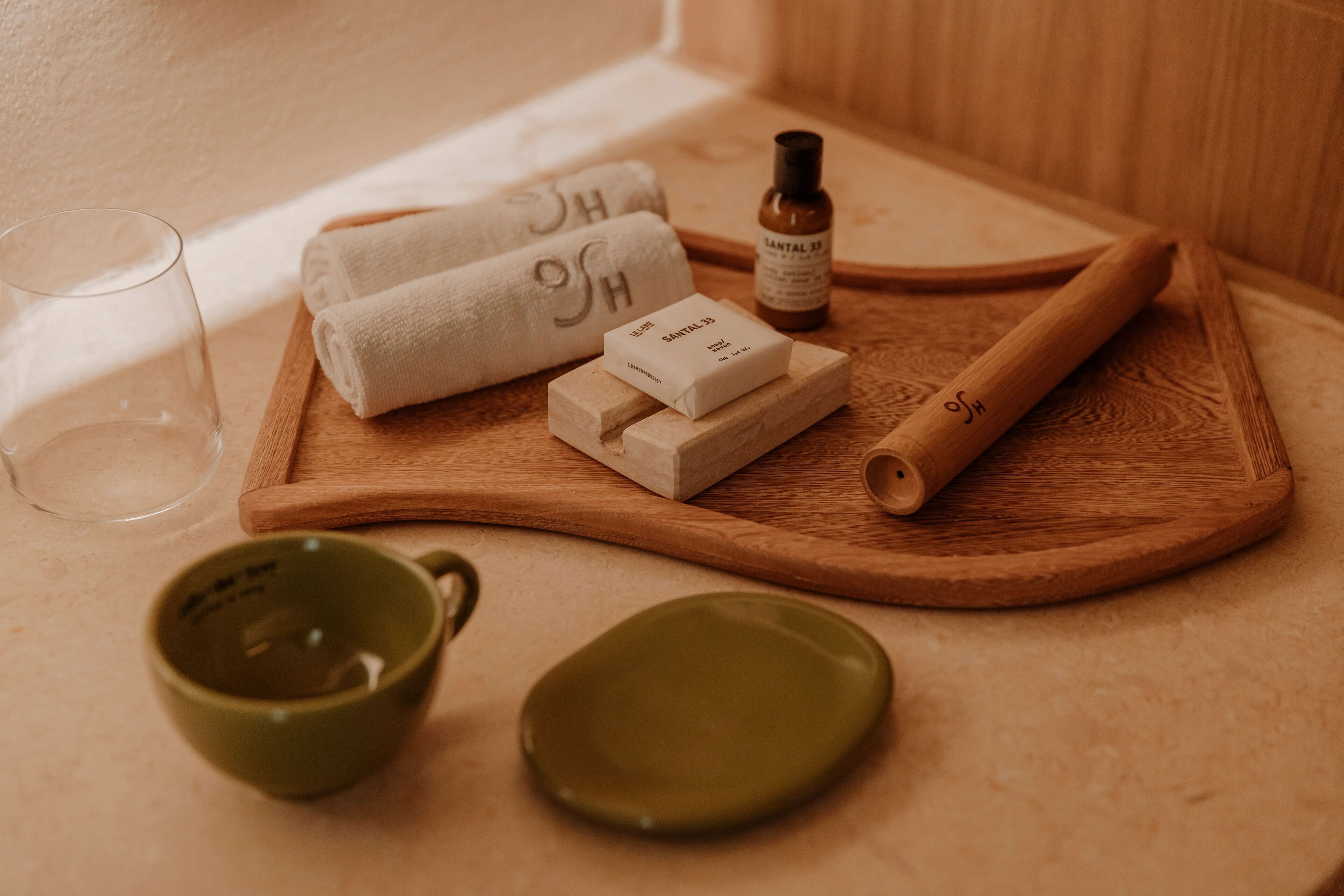 A wooden tray with a bottle of essential oil, a rolled-up towel, a bar of soap, and a wooden rolling pin on a beige countertop.