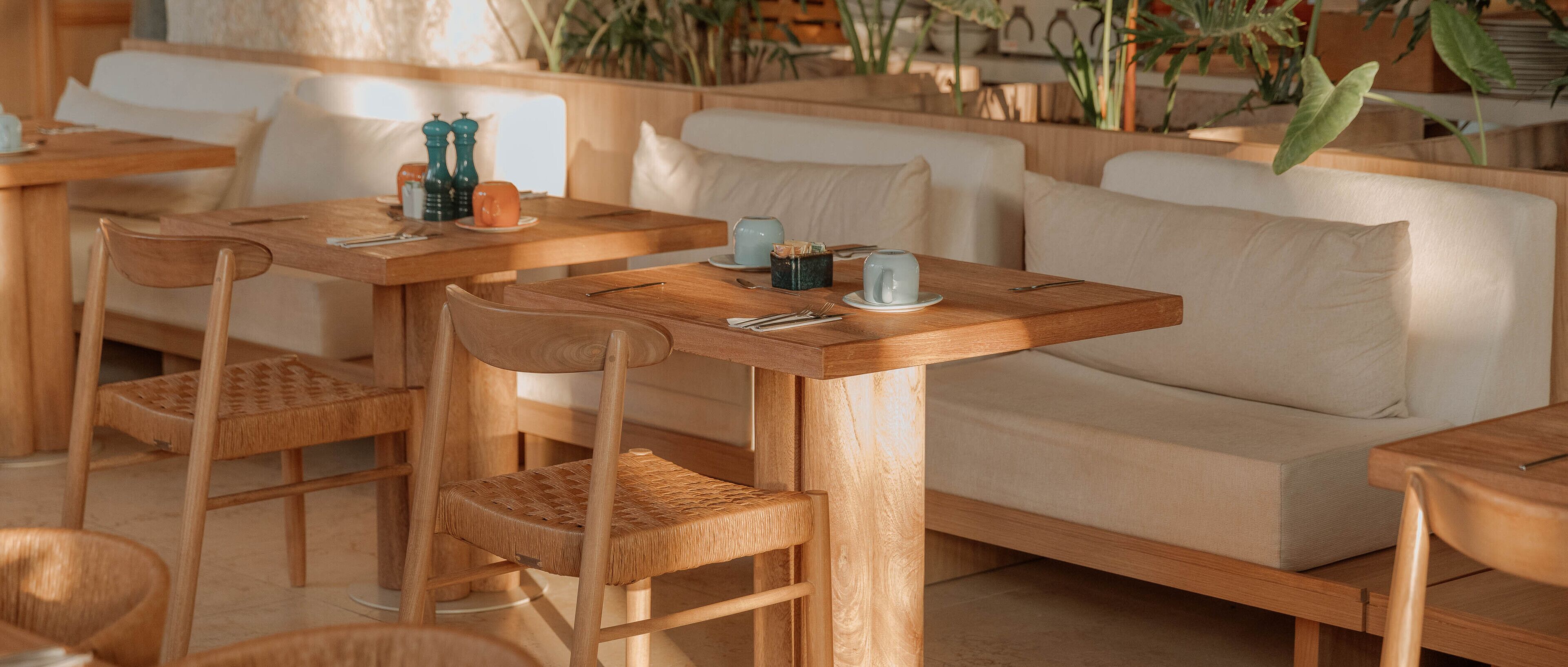 Um café acolhedor com mesas e cadeiras de madeira, um sofá e diversas plantas ao fundo.