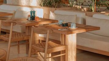Um café acolhedor com mesas e cadeiras de madeira, um sofá e diversas plantas ao fundo.