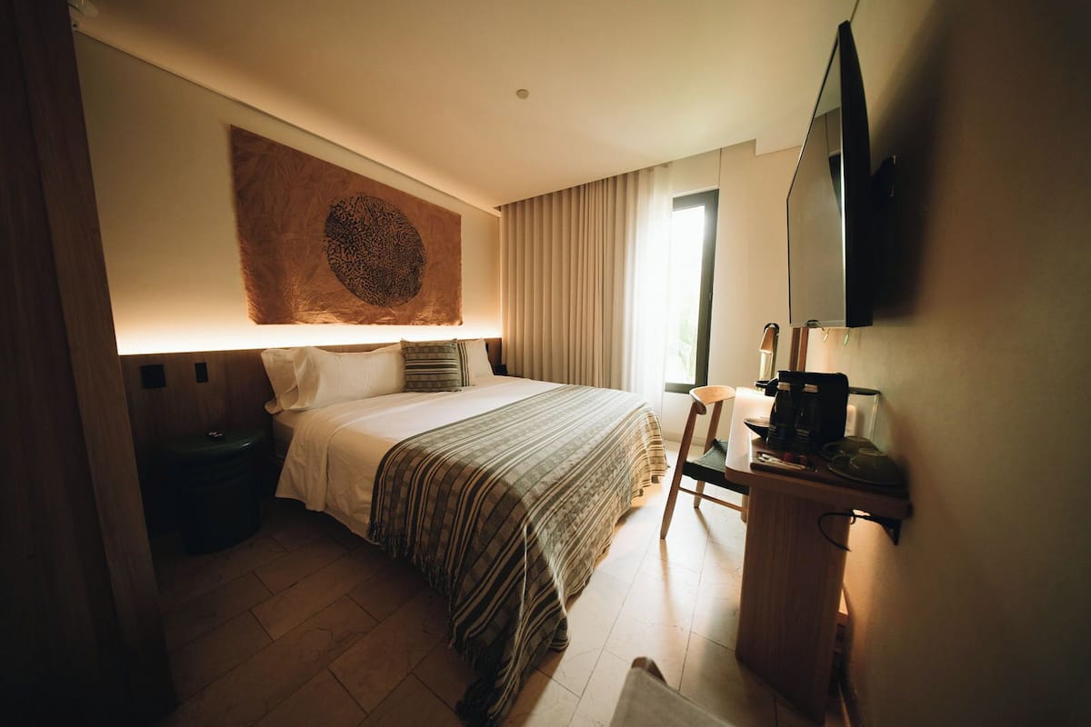 A hotel room with a bed, a desk with a coffee maker, a TV, and a large wall tapestry.