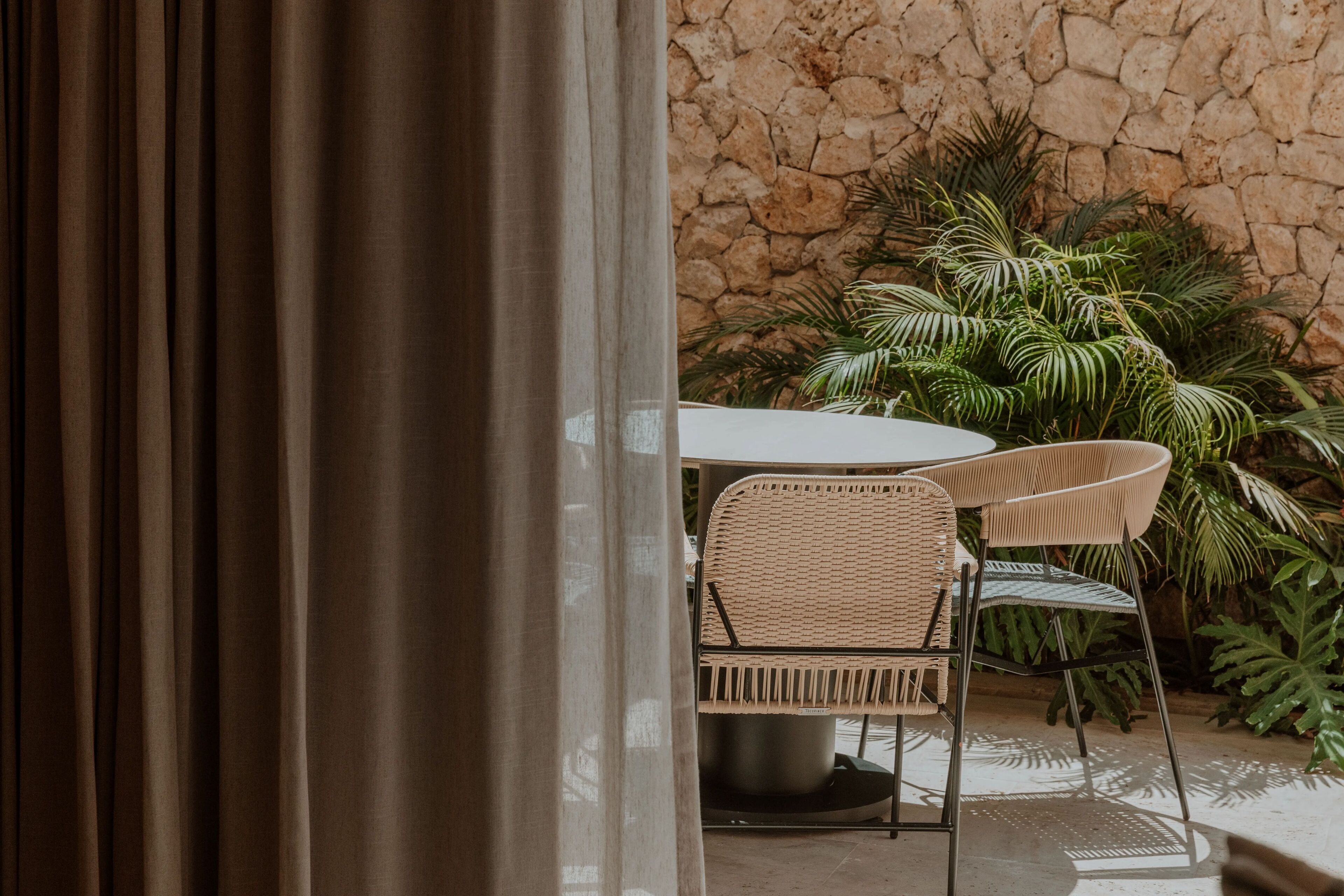 An outdoor seating area with wicker chairs and a table, set against a stone wall and surrounded by lush greenery.