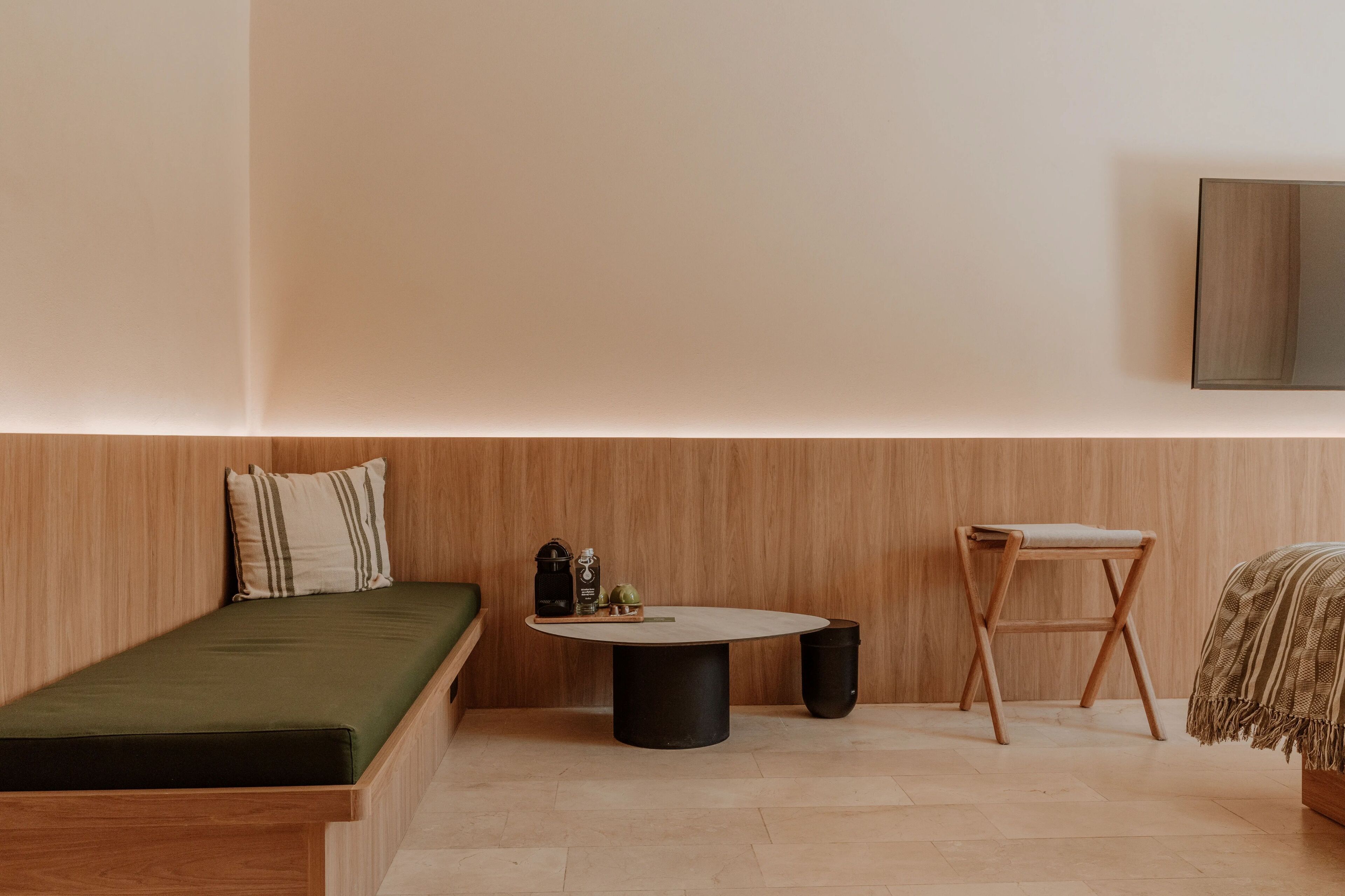 A minimalist room with a wooden wall, a green sofa, a round table with a black base, a wooden stool, and a bed with a striped blanket.