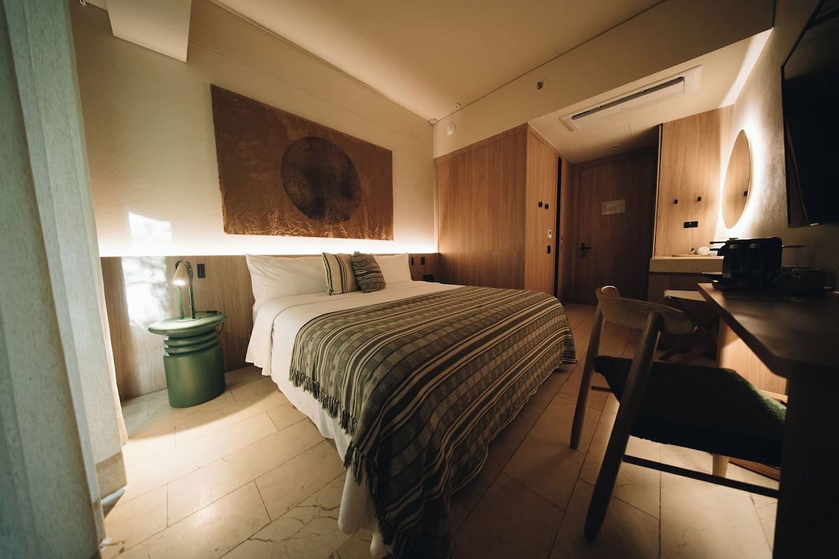 A compact hotel room with a bed, a desk, a chair, a small trash bin, and a wall-mounted artwork.