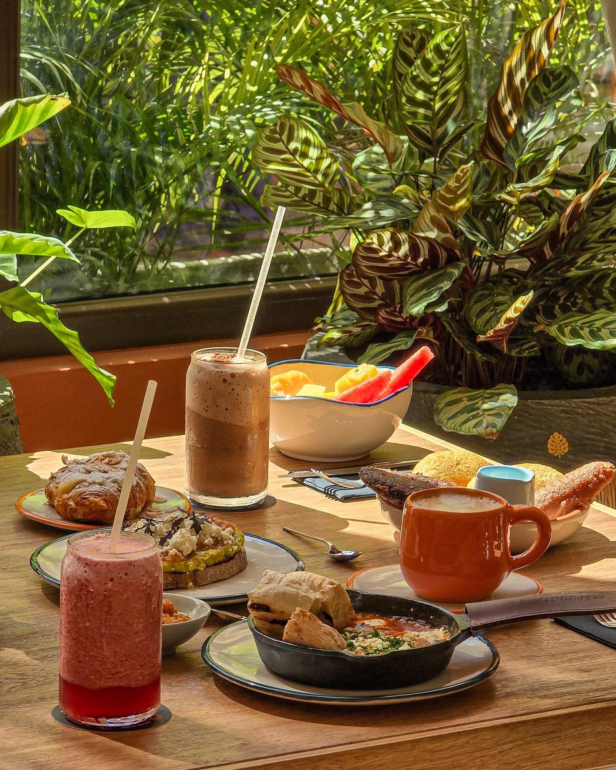 Een ontbijttafel met diverse etenswaren en drankjes, op de achtergrond weelderige groene planten.