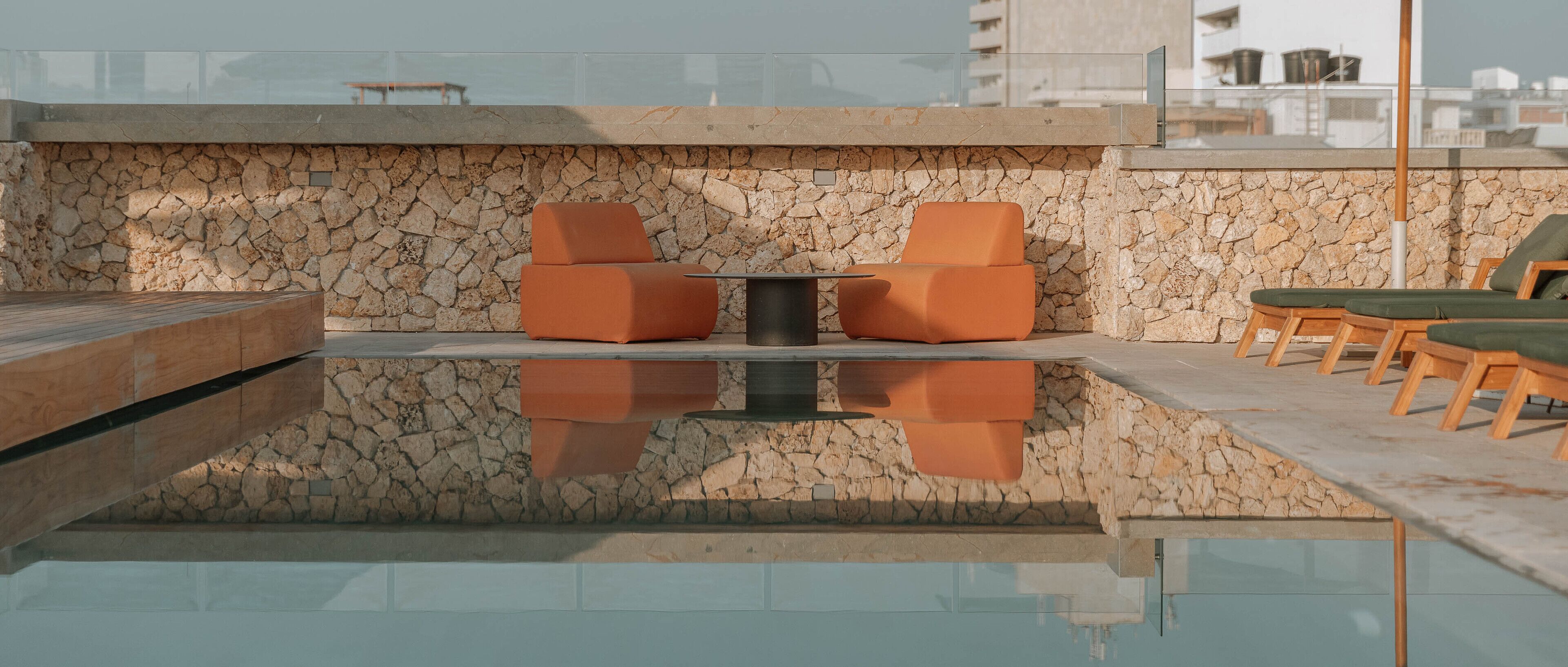 A rooftop pool area with stone walls, orange seating, and a clear view of the cityscape.
