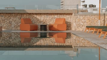 A rooftop pool area with stone walls, orange seating, and a clear view of the cityscape.
