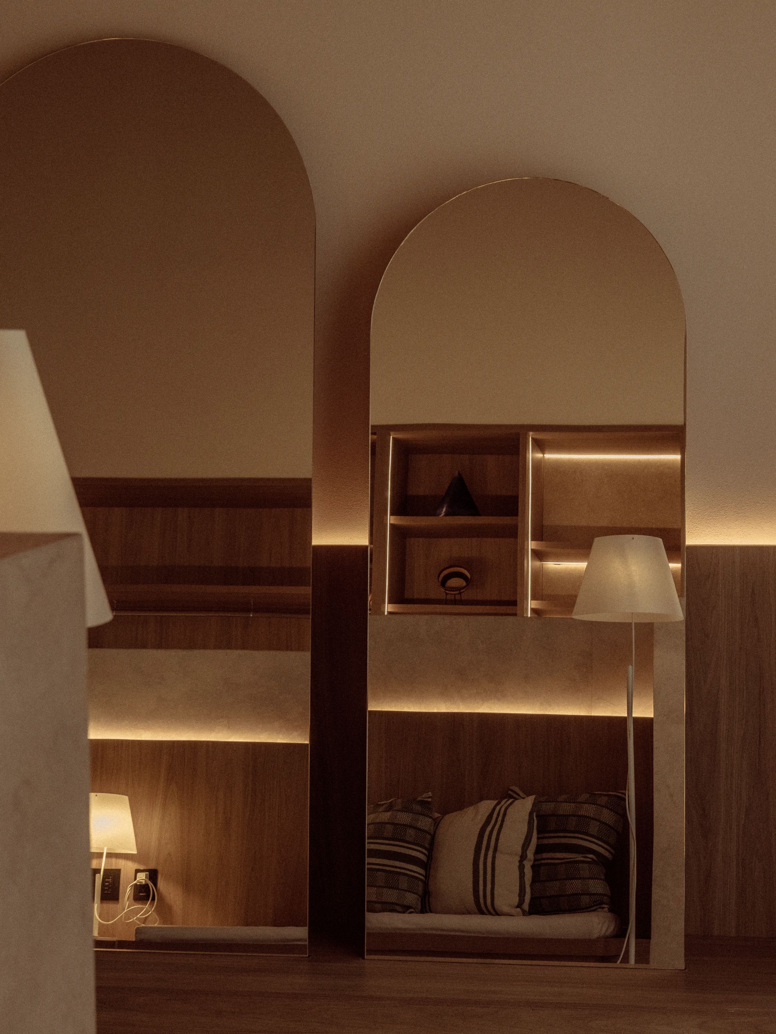 Quarto de hotel moderno com piso de madeira, sofá com almofadas listradas, mesa de cabeceira com abajur e prateleira embutida com luminária.