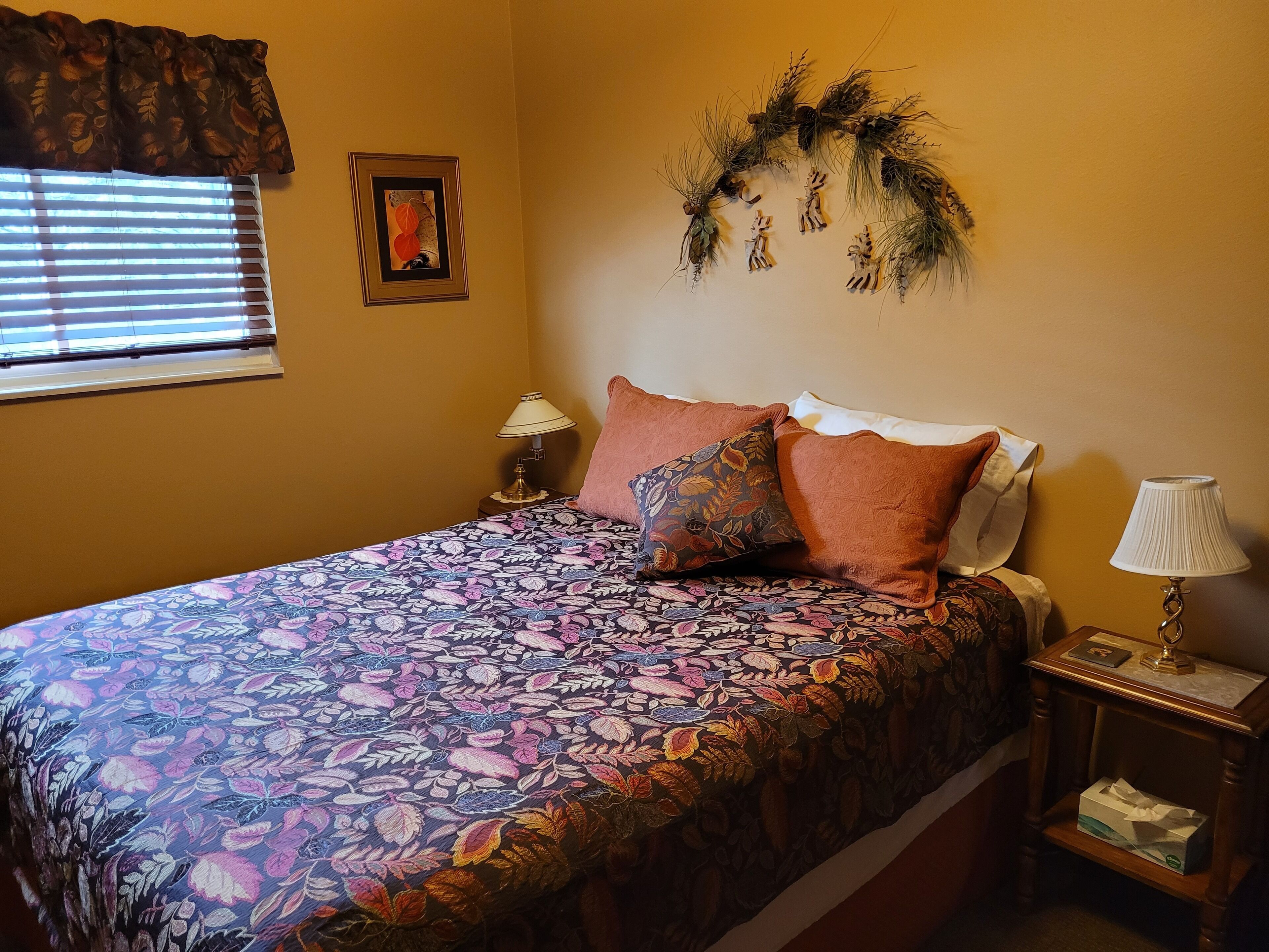 A bedroom with a floral bedspread, two pillows, a nightstand with a lamp, a framed picture, and a wall decoration.