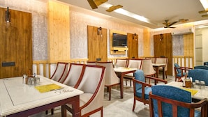 A restaurant with wooden panel walls, multiple tables and chairs, ceiling fans, and a television mounted on the wall. - Holy Shivana Boutique Hotel Rishikesh (Narendranagar)