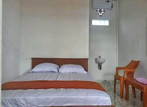 Un lit simple avec une tête de lit en bois, une table de chevet avec une chaise, et un luminaire fixé au mur.