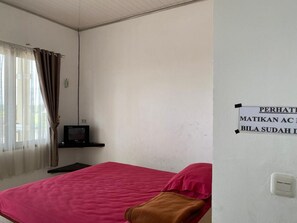 Une chambre avec un lit simple, une couverture rouge, un téléviseur posé sur un meuble et un panneau décoratif au mur.