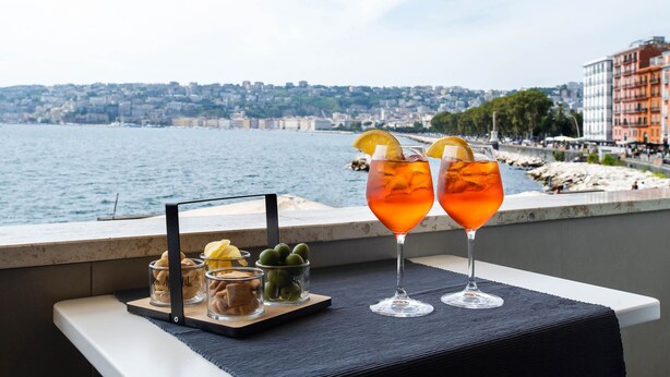 A balcony with two glasses of orange cocktail, a small tray with snacks, and a city view in the background. - Imperatrice Suites (Naples)