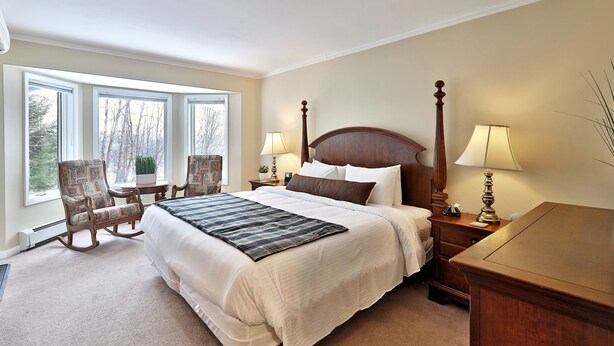 A bedroom with a large bed, two bedside tables with lamps, a chair, and a view of trees through the windows. - The Birch Ridge: English Gentleman's Room #9 - King Suite in Killington, Vermont. Hot Tub. (Killington)
