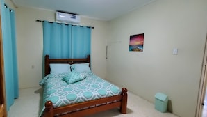 A bedroom with a wooden bed, light blue curtains, a wall-mounted air conditioner, and a small trash bin.