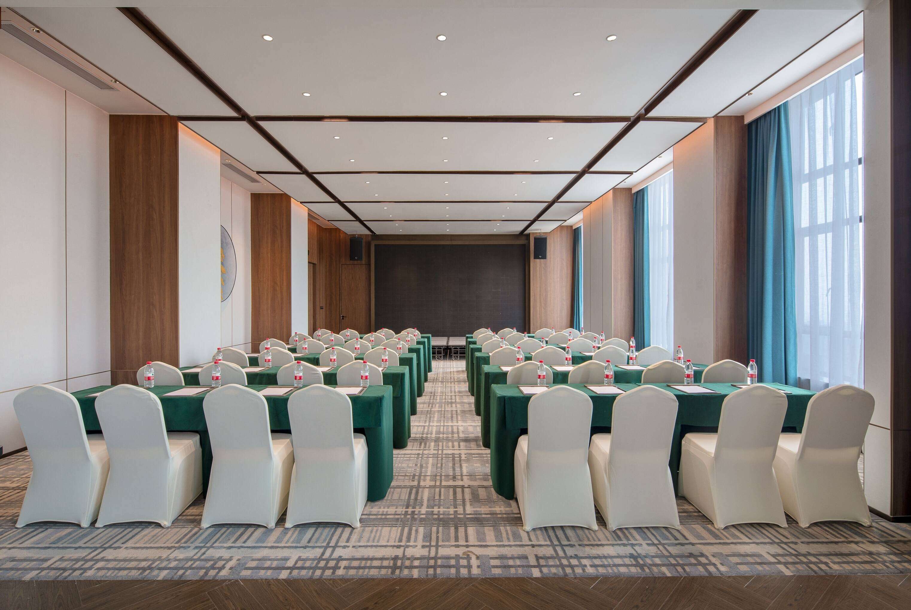 A conference room with rows of tables and chairs, a stage at the front, and large windows with curtains.