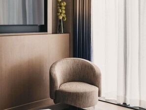 A modern living room with a curved armchair, a flat-screen TV, a vase with yellow flowers, and a window with sheer curtains. - Mondrian Singapore Duxton (Singapore)