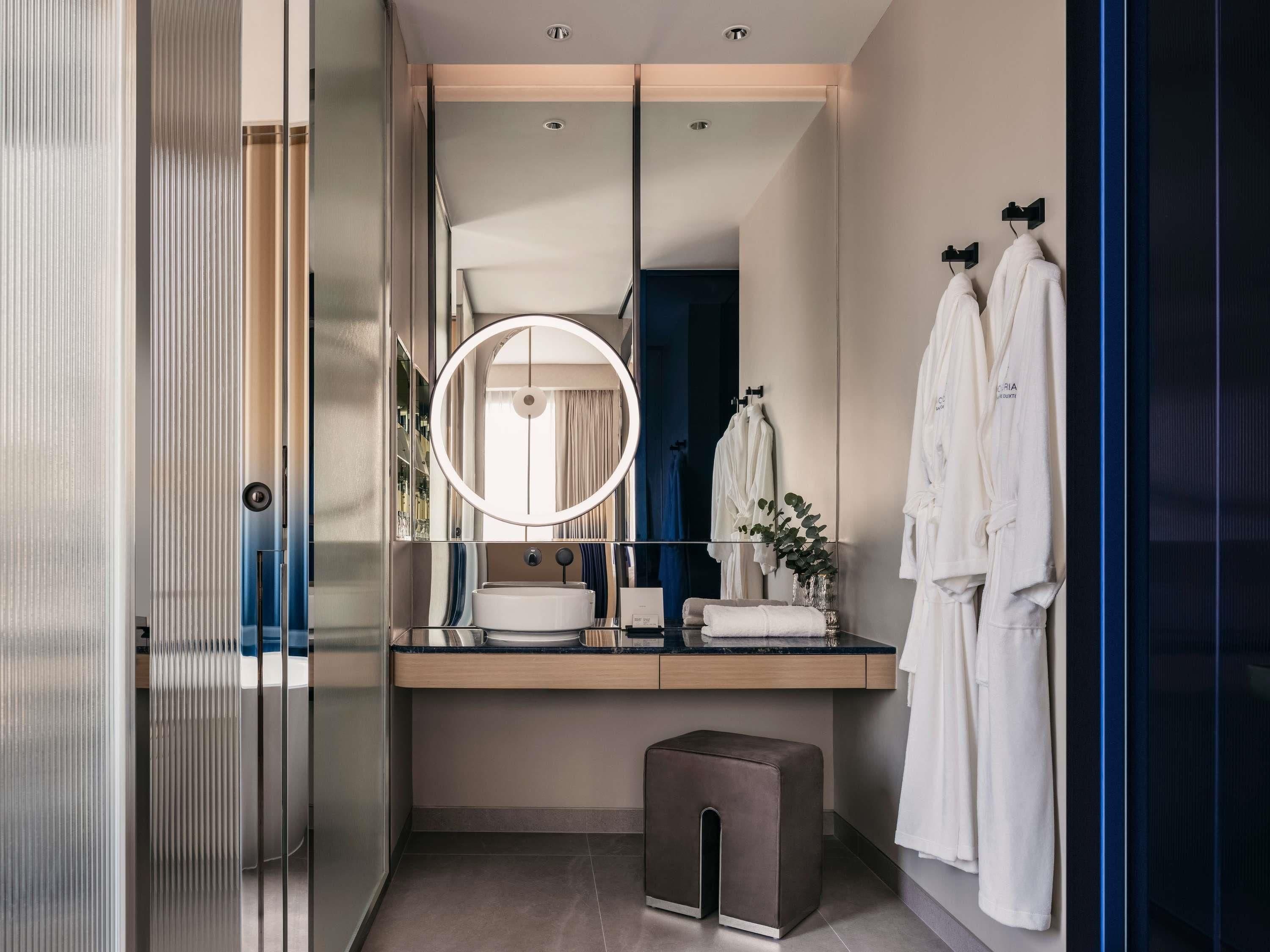 A modern bathroom with a large mirror, a round light, a sink, and two white robes hanging on hooks.