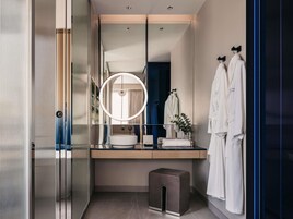 A modern bathroom with a large mirror, a round light, a sink, and two white robes hanging on hooks.