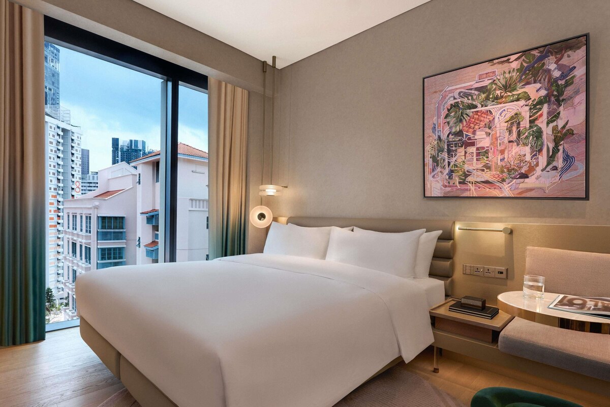 Habitación de hotel con una cama grande, una mesita de noche con lámpara, una pequeña mesa con un vaso de agua, una silla y vistas a la ciudad.
