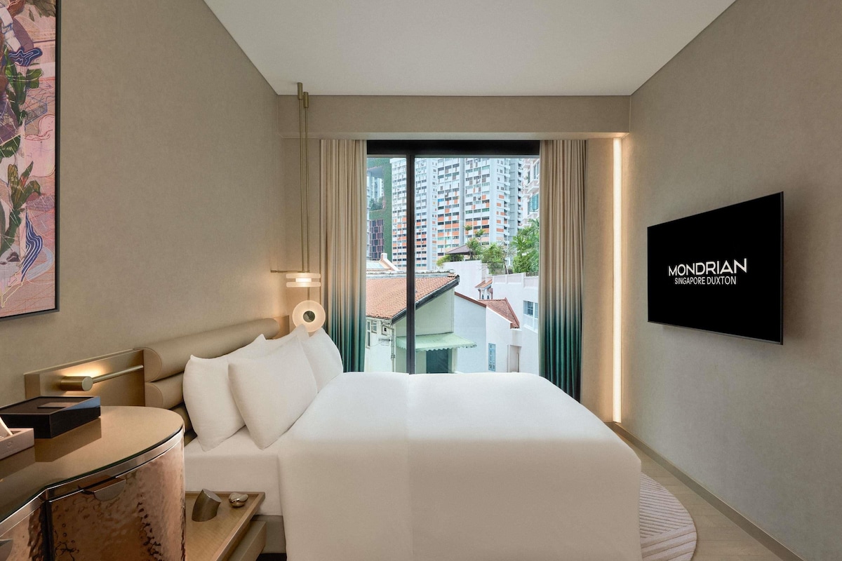 Una habitación de hotel moderna con cama, mesita de noche, lámpara y televisor mostrando el nombre del hotel.