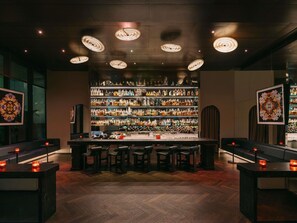 Una zona de bar con suelo de madera tipo espiga, una barra con taburetes y estantes llenos de botellas.