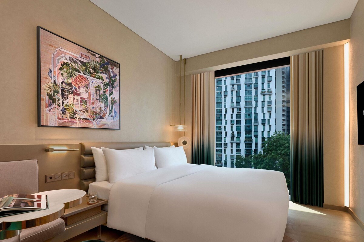Una habitación de hotel moderna con una cama grande, un escritorio con una silla, un ventanal con cortinas y un cuadro en la pared.
