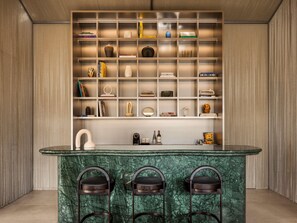 Un área de bar moderna con mostrador de mármol, tres taburetes y una estantería con varios objetos.