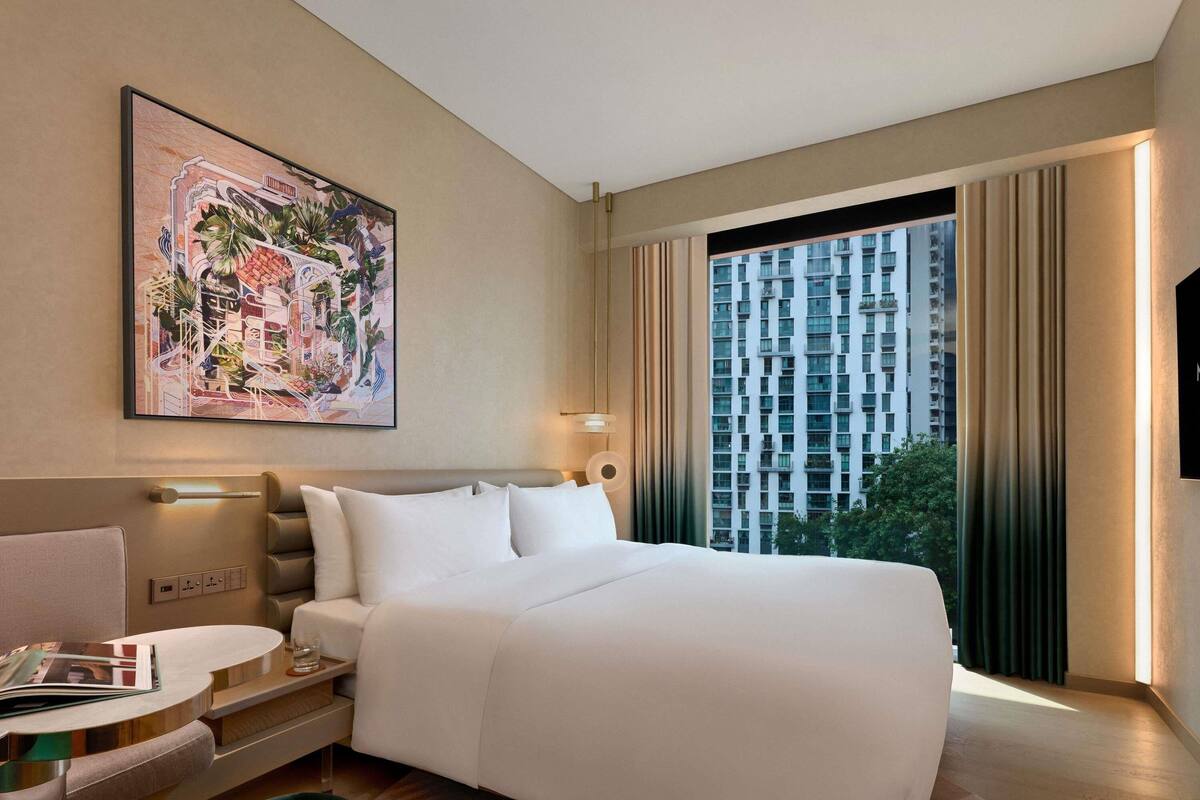 Een moderne hotelkamer met een groot bed, een bureau met stoel, een groot raam met gordijnen en een schilderij aan de muur.