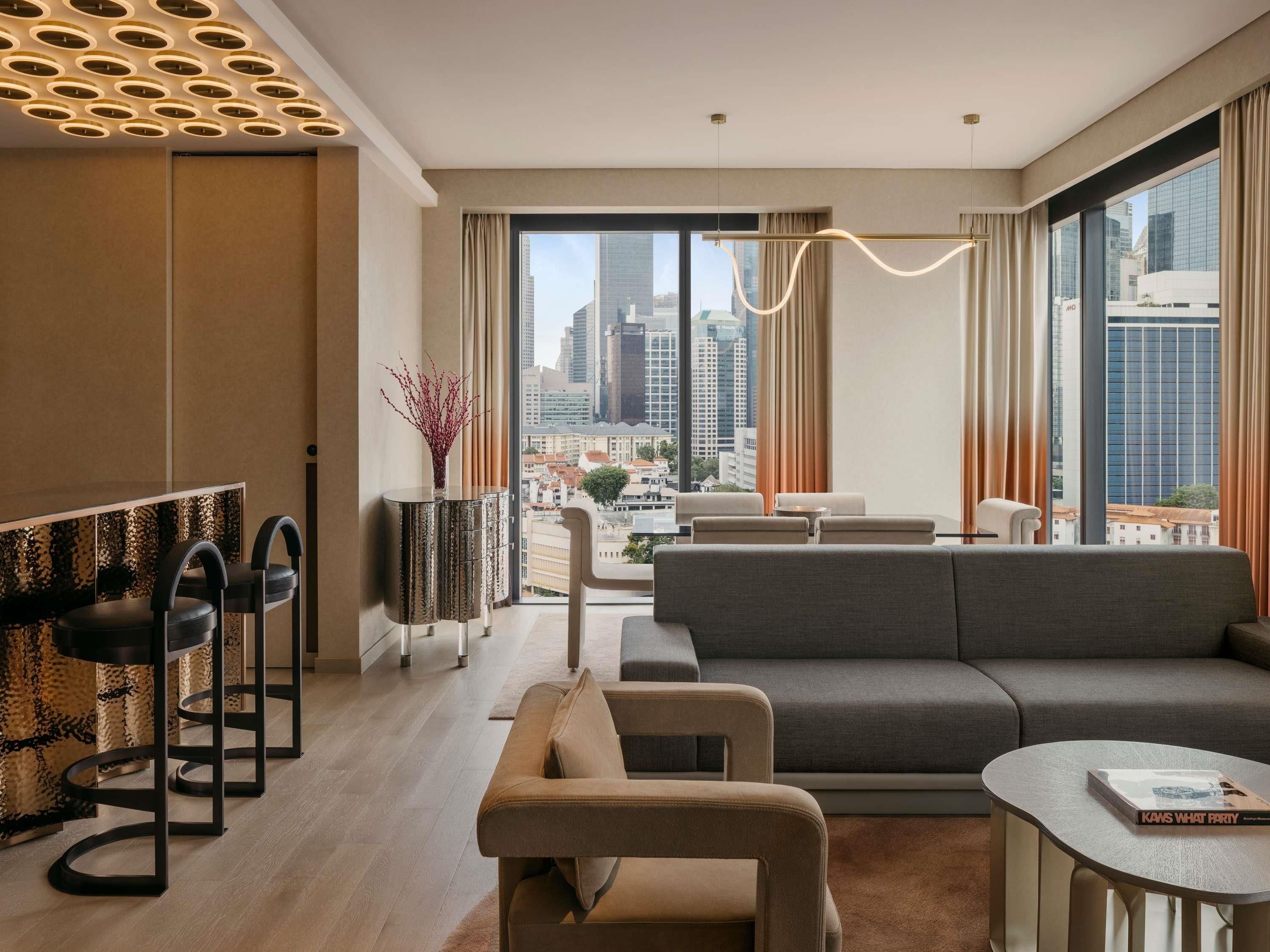 Una habitación de hotel moderna con un ventanal grande, un sofá, una zona de comedor y vistas a la ciudad.