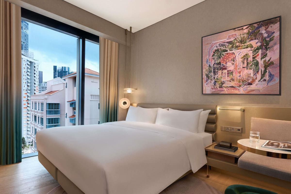 Hotelkamer met een groot bed, nachtkastje met een lamp, een klein tafeltje met een glas water, een stoel en uitzicht over de stad.
