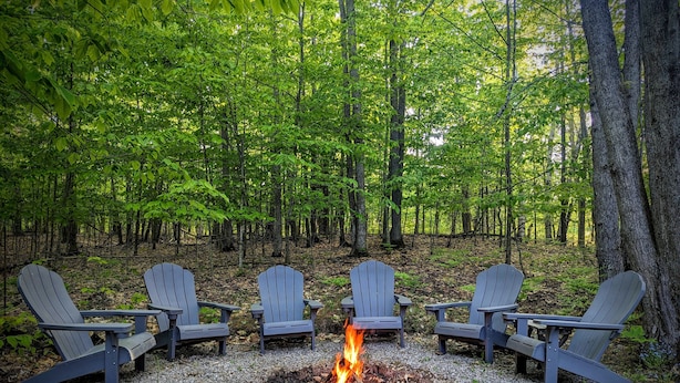 A forest with a fire pit and four Adirondack chairs arranged around it. - Secluded Chalet in Michaywe w/ pool access. Golf + Ski/Snowmobile paradise. (Gaylord)