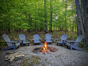 A forest with a fire pit and four Adirondack chairs arranged around it.