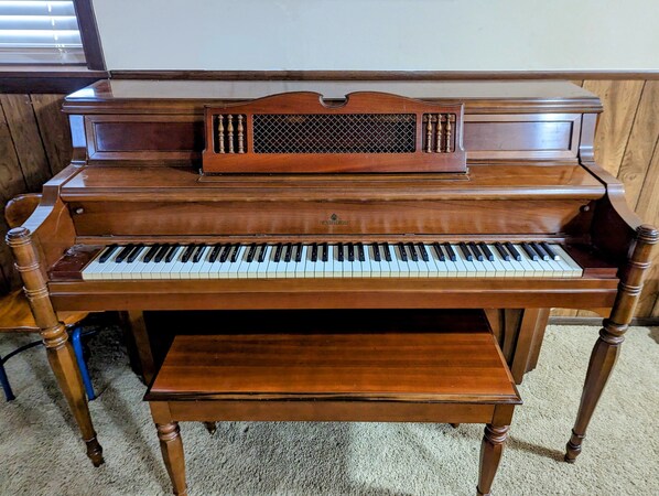 A grand piano with a bench, positioned against a wooden wall with a window in the background.