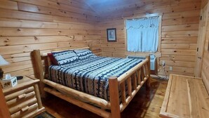 Quarto de cabana de madeira com cama, mesa de cabeceira, janela com cortinas e um quadro na parede.