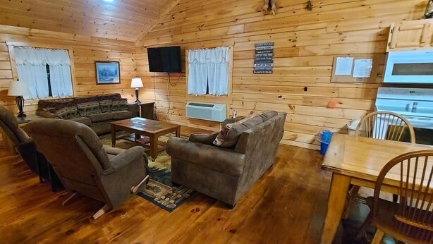 Interior acolhedor de uma cabana com paredes de madeira, um sofá, duas poltronas, uma mesa de centro, uma TV e um espaço com cozinha compacta.