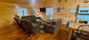 A cozy cabin interior with wooden walls, a sofa, two armchairs, a coffee table, a TV, and a kitchenette area. - Bobcat's Den - Dog Friendly - Sleeps up to 8 (Moravia)